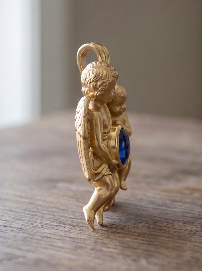 May include: A gold-colored pendant featuring two cherubic figures holding a large, round, blue gemstone. The pendant has a loop at the top for a chain. The cherubs have detailed wings and curly hair.