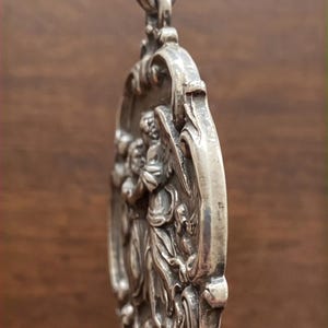 May include: A silver-toned oval pendant featuring a relief sculpture of a winged figure in profile. The pendant has an ornate, decorative border with scrollwork details and a large, integrated bail at the top for hanging on a chain or cord.