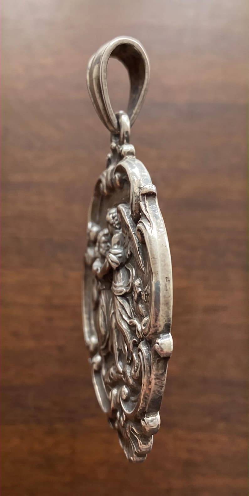 May include: A silver-toned oval pendant featuring a relief sculpture of a winged figure in profile. The pendant has an ornate, decorative border with scrollwork details and a large, integrated bail at the top for hanging on a chain or cord.