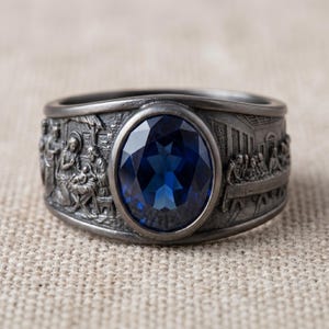 Pode incluir: Um anel prateado com uma grande pedra preciosa oval azul escuro no centro. A banda apresenta entalhes em relevo detalhados de cenas religiosas. O design do anel é intrincado e a pedra preciosa é facetada.