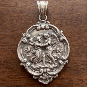 May include: A silver pendant featuring a detailed relief of a person wrestling with an angel, surrounded by two small cherubs. The circular piece has an ornate, scrolled border and a polished silver bail at the top for hanging on a chain.