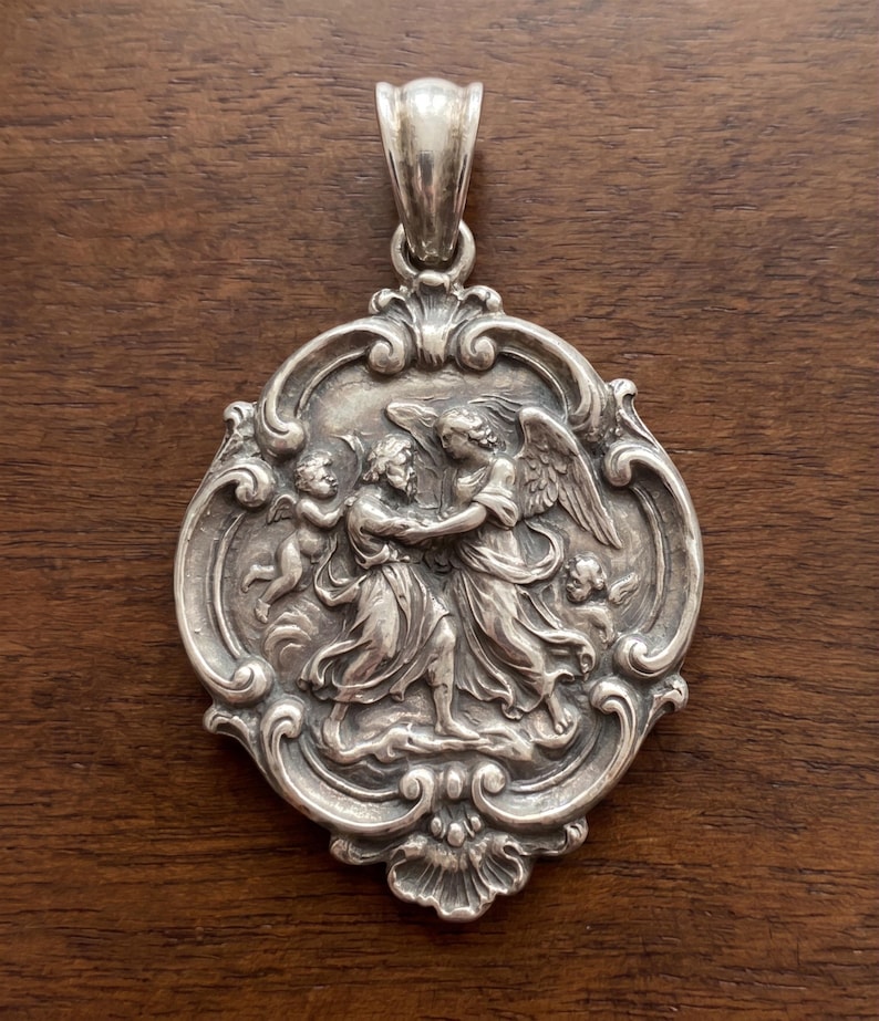 May include: A silver pendant featuring a detailed relief of a person wrestling with an angel, surrounded by two small cherubs. The circular piece has an ornate, scrolled border and a polished silver bail at the top for hanging on a chain.