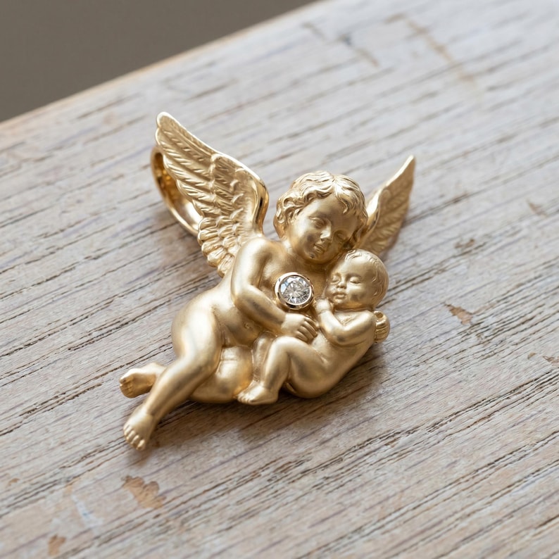 May include: A gold-colored pendant featuring two cherubic figures, one embracing the other. The pendant has detailed wings and a small, round, clear gemstone in the center. The pendant is on a wooden surface.