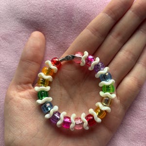 May include: A circular bracelet featuring a repeating pattern of translucent, multi-coloured plastic beads separated by small, white, wavy-shaped spacers. The beads include shades of red, orange, yellow, green, blue, purple, and pink.