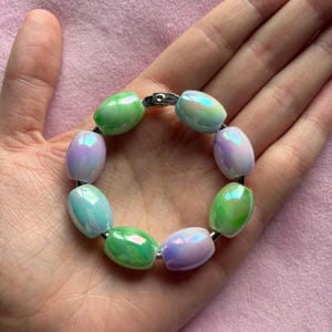 Peut inclure: Un bracelet circulaire composé de perles en forme de tonneau alternées avec une finition irisée dans des tons de vert pastel, bleu clair et lavande doux. Les perles sont enfilées sur un fil métallique argenté fixé par un petit fermoir.