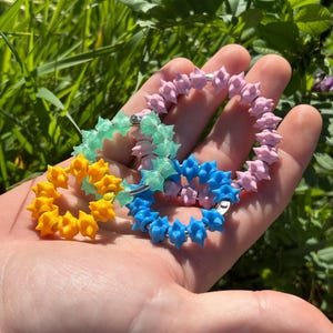 Peut inclure: Quatre anneaux circulaires ornés de petites perles en forme d'étoile aux couleurs vives. L'ensemble comprend un anneau avec des perles rose clair, un jaune vif, un vert menthe translucide et un bleu uni sur des anneaux métalliques.