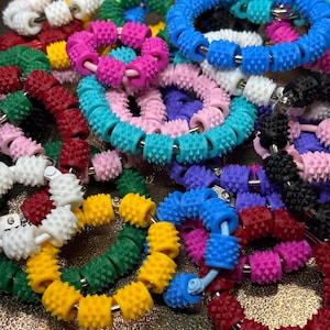 May include: A collection of circular sensory rings featuring small, textured, spiky silicone beads in various vibrant colours including yellow, white, green, pink, blue, purple, red, and black, all strung together on a flexible metal wire frame.