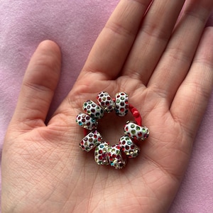 May include: A red elastic hair tie adorned with multiple silver-toned cylindrical beads. Each bead features a white enamel finish and is encrusted with small, multicolored rhinestones in shades of red, green, blue, and clear, creating a vibrant, circular accessory.
