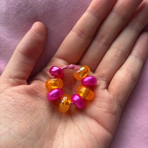 May include: A circular pink metal ring featuring a series of alternating translucent orange and opaque metallic pink beads threaded onto the band. The beads are smooth and rounded, creating a vibrant, colourful contrast against the pink metal frame.