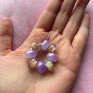 Op de afbeelding: Een cirkelvormig haaraccessoire met afwisselend gladde lavendelkleurige kralen en doorschijnende kralen met gouden glitterspikkels, geregen aan een flexibele, lichtpaarse elastische band die een kleine ringvorm vormt.