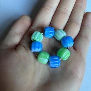 Pode incluir: Uma pulseira com oito contas de vidro coloridas em tons de azul e verde, enfiadas em um fio de prata. As contas têm uma textura canelada e uma aparência marmorizada, criando um design único e atraente.