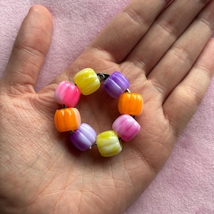 May include: A circular metal ring adorned with seven translucent, ribbed, barrel-shaped beads in vibrant shades of yellow, purple, orange, and pink. The beads are arranged in a continuous loop around the thin silver-toned metal band.
