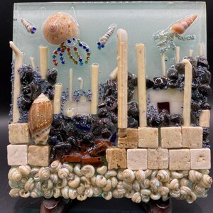 以下が含まれることがあります： 混合素材を使用した水中シーンを描いた正方形のガラスアート作品。カラフルなラインストーンの触手を持つ螺旋状の貝殻のクラゲ、小さな貝殻、黒い石、白い石の柱、そして多くの小さな白い貝殻で構成された土台が特徴です。