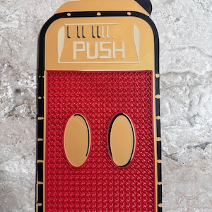 May include: A red, black, and gold decorative push button with a Mickey Mouse theme. The button has a textured red center, two gold ovals, and the word "PUSH" in white.