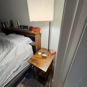 May include: A dark wood bedside table with two tiers and a black metal frame, featuring a tall floor lamp with a cylindrical beige fabric shade attached to the back. The table surface holds a white charging puck, a pen, and a small gold case.