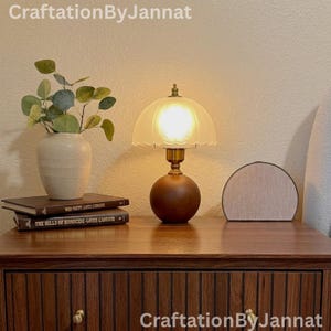 May include: A vintage-style table lamp with a frosted glass shade and a dark brown wooden base. The lamp is on a wooden nightstand with a vase of greenery, books, and a speaker. The text "CraftationByJannat" is visible.