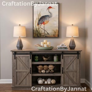 May include: A rustic wooden cabinet with sliding barn doors, flanked by two gray geometric lamps with beige shades. Above, a framed painting of a heron stands out. Decorative items include candles, a bird figurine, and woven spheres.