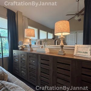 May include: A long, dark brown wooden sideboard with horizontal slatted cabinet doors. Two matching table lamps with light-colored, textured fabric shades and turned wooden bases sit on top. A white picture frame and a small vase are also present.
