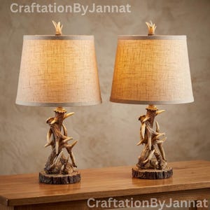 May include: A pair of table lamps featuring bases constructed from stacked, textured brown antlers resting on circular wood-grain platforms. Each lamp is topped with a tapered, light tan fabric shade and a small, pointed finial at the top.