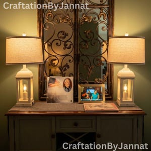 May include: Two white lantern-style table lamps with beige fabric drum shades flank a central decorative arrangement. The lamps feature a rectangular base with a visible light bulb inside. The text CraftationByJannat appears at the top and bottom.