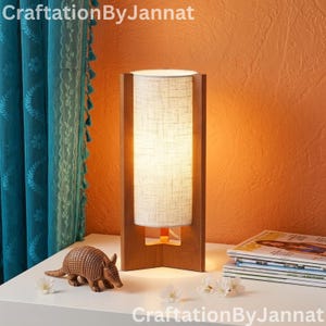 May include: A wooden table lamp with a rectangular frame and a cylindrical, off-white fabric shade. The lamp is on a white surface, next to a small armadillo figurine and a stack of magazines. The background is orange.
