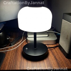 May include: A table lamp with a matte black circular base and a thin black stem supporting a large, glowing white dome-shaped shade. The lamp emits a bright, even light. The text CraftationByJannat is visible at the top and bottom of the image.