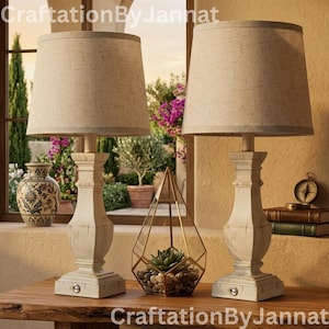 May include: Two cream-colored table lamps with beige shades sit on a wooden table. The lamps have a distressed white base. A geometric terrarium with a succulent and a small compass are also on the table. The text "CraftationByJannat" is visible.