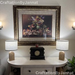 May include: A still life painting of fruits and flowers in a gold frame hangs above a white table. Two lamps with beige shades and wooden bases flank a black clock. The text "CraftationByJannat" is visible.