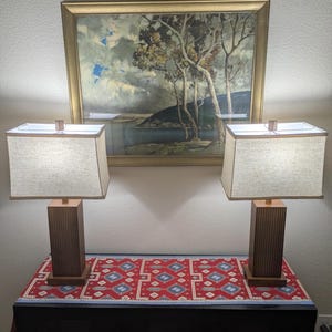 May include: Two wooden table lamps with rectangular shades sit on a table with a red and blue patterned runner. A framed landscape painting hangs above the table. The lamps have a square base and a light-colored shade.