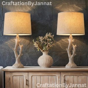 May include: Two table lamps with beige fabric drum shades and light-colored bases shaped like tree branches. Between the lamps sits a round, ribbed ceramic vase containing dried flowers. The text CraftationByJannat appears in the image.