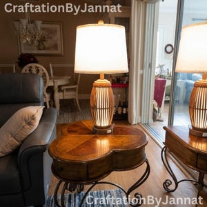 May include: A table lamp with a cylindrical slatted wooden base and a white fabric drum shade, resting on a clover-shaped wooden side table with curved metal legs. The lamp emits a warm glow and features a small power switch on the base.