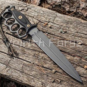 May include: A matte black fixed-blade knife featuring a knuckle duster handle with four finger holes, a textured grip secured by three brass-coloured screws, a double-edged spear point blade, and a thin cord lanyard attached to the pommel end.