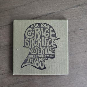 May include: A square canvas print with a pale green background. The print features a silhouette of a military helmet filled with the words "FOR YOUR COURAGE SACRIFICE & SERVICE WE THANK YOU" in dark grey.