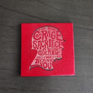 May include: A red square with a textured surface features a white silhouette of a person's head wearing a hat. The text inside the silhouette reads "For Your Courage, Sacrifice & Serve, We Thank You."