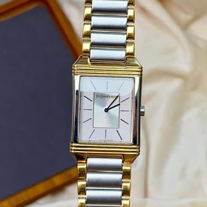 May include: A rectangular wristwatch with a two-tone gold and silver metal link bracelet. The watch face is white with thin black hour markers, black hands, and the text Yves Saint Laurent printed in black near the top center of the dial.