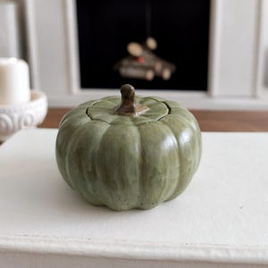 May include: A sage green ceramic pumpkin-shaped lidded container with a smooth, glossy finish and a dark brown stem handle. The pumpkin features vertical ridges and a rounded, squat silhouette, providing a decorative storage solution.