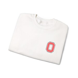 Può includere: Felpa color crema con una toppa rossa e nera a forma di lettera "O" sul petto. La felpa è piegata ordinatamente, mettendo in risalto lo scollo tondo. Uno stile semplice e classico.