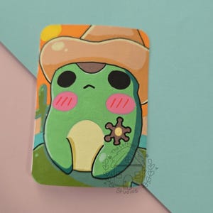May include: A rectangular sticker featuring a green frog wearing a tan cowboy hat. The frog has large black eyes, pink cheeks, and a brown sheriff star badge. The background shows a desert landscape with a yellow sun and a cactus. The text The Cottage Studios is visible.