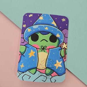 May include: A rectangular sticker featuring a green frog character wearing a blue wizard hat and cape decorated with yellow stars. The frog has black eyes, pink cheeks, and holds a small yellow star wand. The text The Cottage Fox Studio is visible.