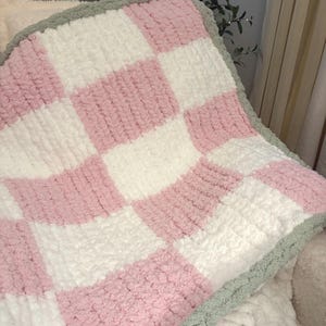 May include: A chunky knit blanket featuring a checkered pattern of soft pink and white squares. The blanket is finished with a textured sage green border that frames the entire piece, showcasing a thick, plush yarn construction throughout.