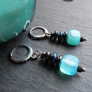 May include: A pair of dangle earrings with a light blue, square bead. The earrings have a silver clasp and are accented with dark blue and silver beads. The earrings are on a dark grey surface.