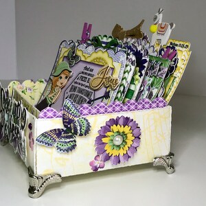 K&ouml;nnte beinhalten: Eine wei&szlig;e Box mit einem floralen Design in Lila und Gelb. Die Box hat einen gewellten Rand und ist mit Schmetterlingen, Blumen und einem M&auml;dchen mit einem gr&uuml;nen Hut verziert. Die Box ist mit Karten gef&uuml;llt und hat auf einer der Karten ein Zitat &uuml;ber Jesus. Die Box steht auf einer wei&szlig;en Oberfl&auml;che.