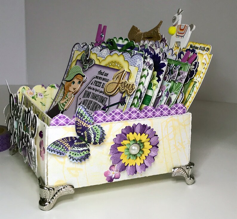 K&ouml;nnte beinhalten: Eine wei&szlig;e Box mit einem floralen Design in Lila und Gelb. Die Box hat einen gewellten Rand und ist mit Schmetterlingen, Blumen und einem M&auml;dchen mit einem gr&uuml;nen Hut verziert. Die Box ist mit Karten gef&uuml;llt und hat auf einer der Karten ein Zitat &uuml;ber Jesus. Die Box steht auf einer wei&szlig;en Oberfl&auml;che.