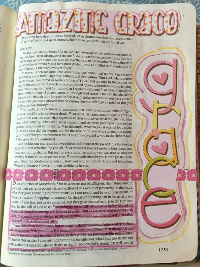 May include: A page from a book featuring the text Amazing Grace in floral-patterned letters at the top and the word Grace in pink and yellow vertical letters with heart accents on the right side, alongside pink decorative tape and text.