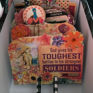 May include: A scrapbook page with an autumn theme. The page features a quote about God giving his toughest battles to his strongest soldiers. The page is decorated with autumn-themed embellishments, including a turkey, flowers, and leaves.