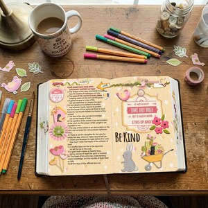 May include: An open book with printed text and decorative stickers featuring a rabbit, flowers, and a wheelbarrow, surrounded by colored pens, a mug, a jar of buttons, scissors, and paper cutouts on a wooden surface. The text reads Be Kind.