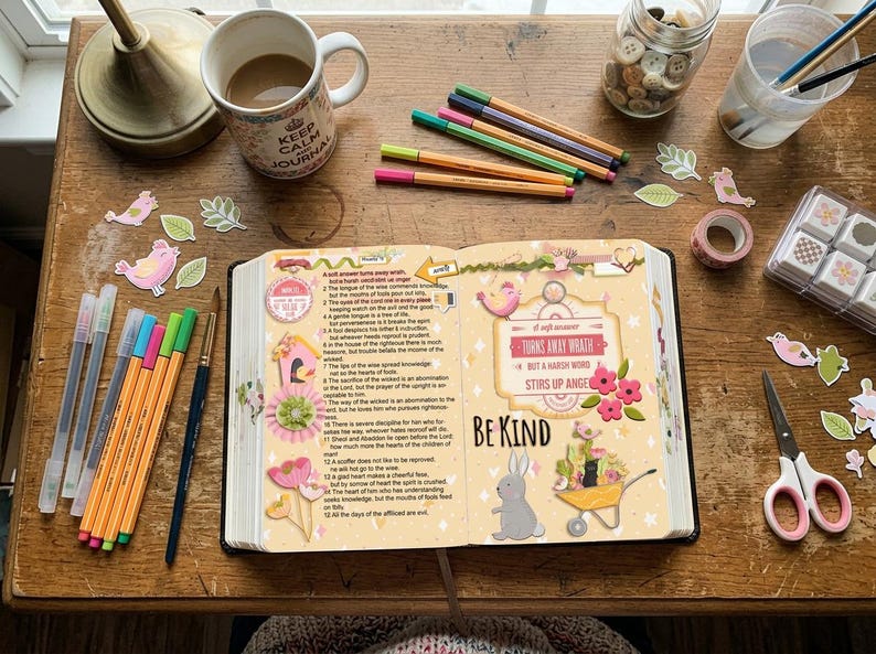 May include: An open book with printed text and decorative stickers featuring a rabbit, flowers, and a wheelbarrow, surrounded by colored pens, a mug, a jar of buttons, scissors, and paper cutouts on a wooden surface. The text reads Be Kind.