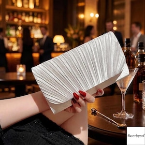 May include: A silver pleated clutch bag with a rectangular shape. The clutch is held in front of a bar setting with a martini glass and bottles in the background. The clutch has a textured, fan-like design.