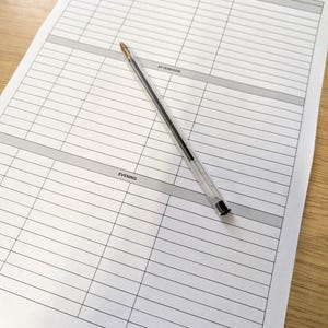 Könnte beinhalten: Ein weißes Papierplaner mit Rasterlayout für die tägliche Planung, mit Abschnitten für Morgen, Nachmittag und Abend. Ein durchsichtiger Kunststoffstift mit schwarzer Tinte liegt diagonal über dem Planer.