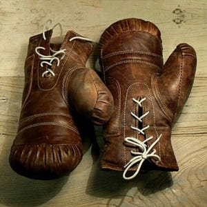 Puede incluir: Un par de guantes de boxeo de cuero marrón de estilo vintage con acabado desgastado, costuras reforzadas y cierres de cordones blancos en la parte delantera. Los guantes tienen una sección de pulgar redondeada y un diseño de puño fruncido.